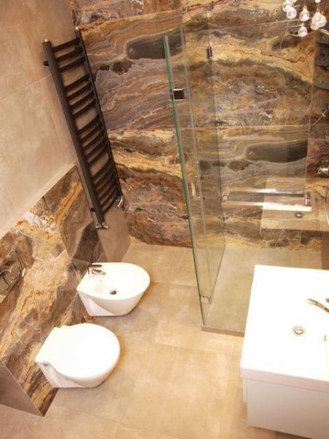 Wnętrze łazienki z marmurowymi ścianami, kabiną prysznicową ze szkła, grzejnikiem drabinkowym, bidetem i umywalką, oświetlone dekoracyjnym żyrandolem.