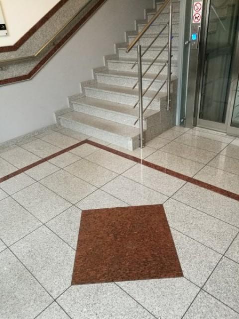 Schody kamienne z jasnego granitu z metalową balustradą, widoczne płytki podłogowe z ciemniejszym kwadratem, obok winda.