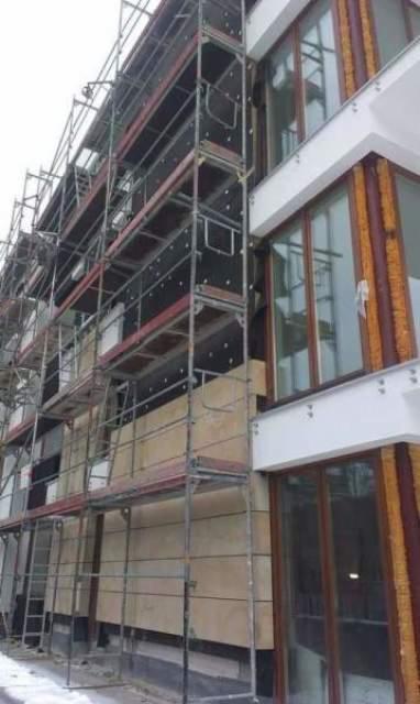 Budynek w trakcie ocieplania, widoczne rusztowania i płyty izolacyjne na elewacji, okna z brązowymi ramami.
