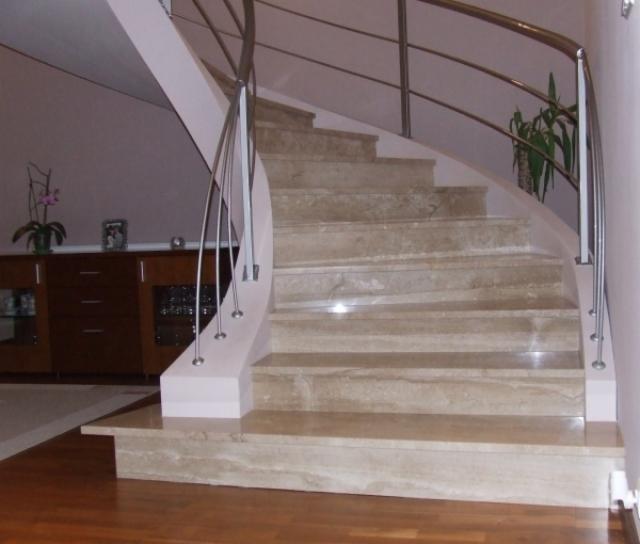 Spiralne schody z trawertynu z metalową balustradą, widok z dołu, drewniana podłoga, w tle komoda z roślinami doniczkowymi.