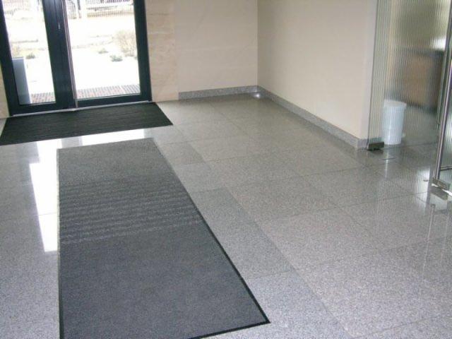 Granitowa posadzka w holu wejściowym, widoczne dwa wycieraczki pod drzwiami, szara kolorystyka.