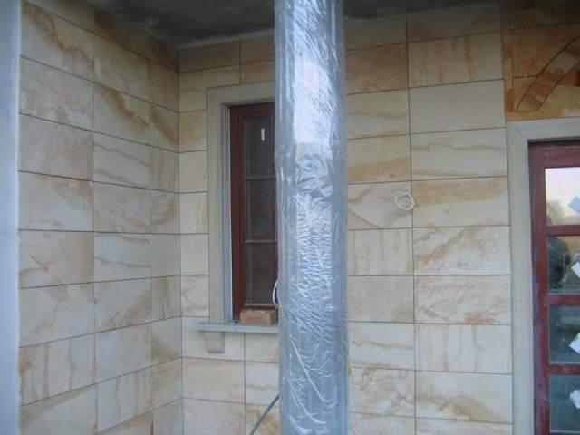 Elewacja budynku wykończona płytami z piaskowca, widoczna kolumna osłonięta folią ochronną, okno z brązową ramą i fragment drzwi.