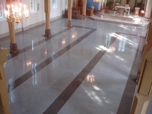 Polerowana posadzka z szarego kamienia z ciemnobrązowymi pasami wzdłuż nawy kościoła, odbijająca światło z okien i żyrandoli.