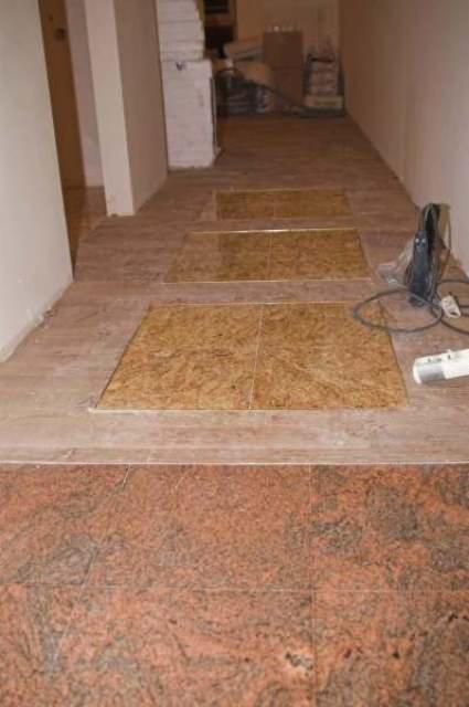 Fragment korytarza z częściowo ułożoną podłogą z płytek w różnych odcieniach brązu i beżu, widoczne narzędzia i materiały budowlane.