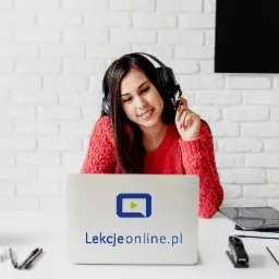 Uśmiechnięta kobieta w słuchawkach przed laptopem z logo lekcjeonline.pl, biała ceglana ściana w tle, minimalistyczne biurko z sukulentami, tablet i notatnik obok.