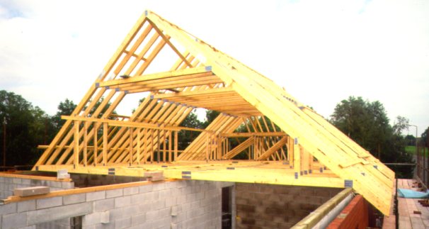 Wznoszenie drewnianej konstrukcji dachu z widocznymi wiązarami na murowanym budynku w trakcie budowy, z zielenią w tle.