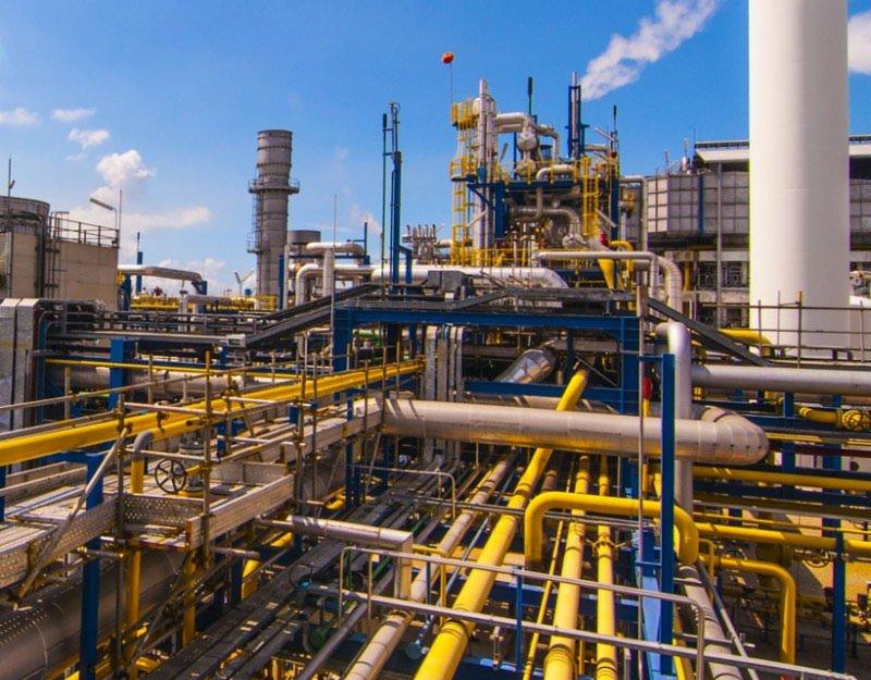 Rozległa instalacja przemysłowa z rurami w kolorach żółtym i szarym, podpartymi niebieską konstrukcją, z kominami i zbiornikami na tle błękitnego nieba z lekkimi obłokami.