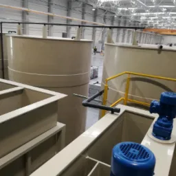 Instalacja przemysłowa z dużymi zbiornikami i niebieskimi pompami w jasnej hali produkcyjnej z widoczną konstrukcją dachu.