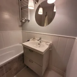 Jasna łazienka z owalnym lustrem, umywalką z szafką na nóżkach, grzejnikiem drabinkowym i wanną z dekoracyjną mozaiką przy obudowie.