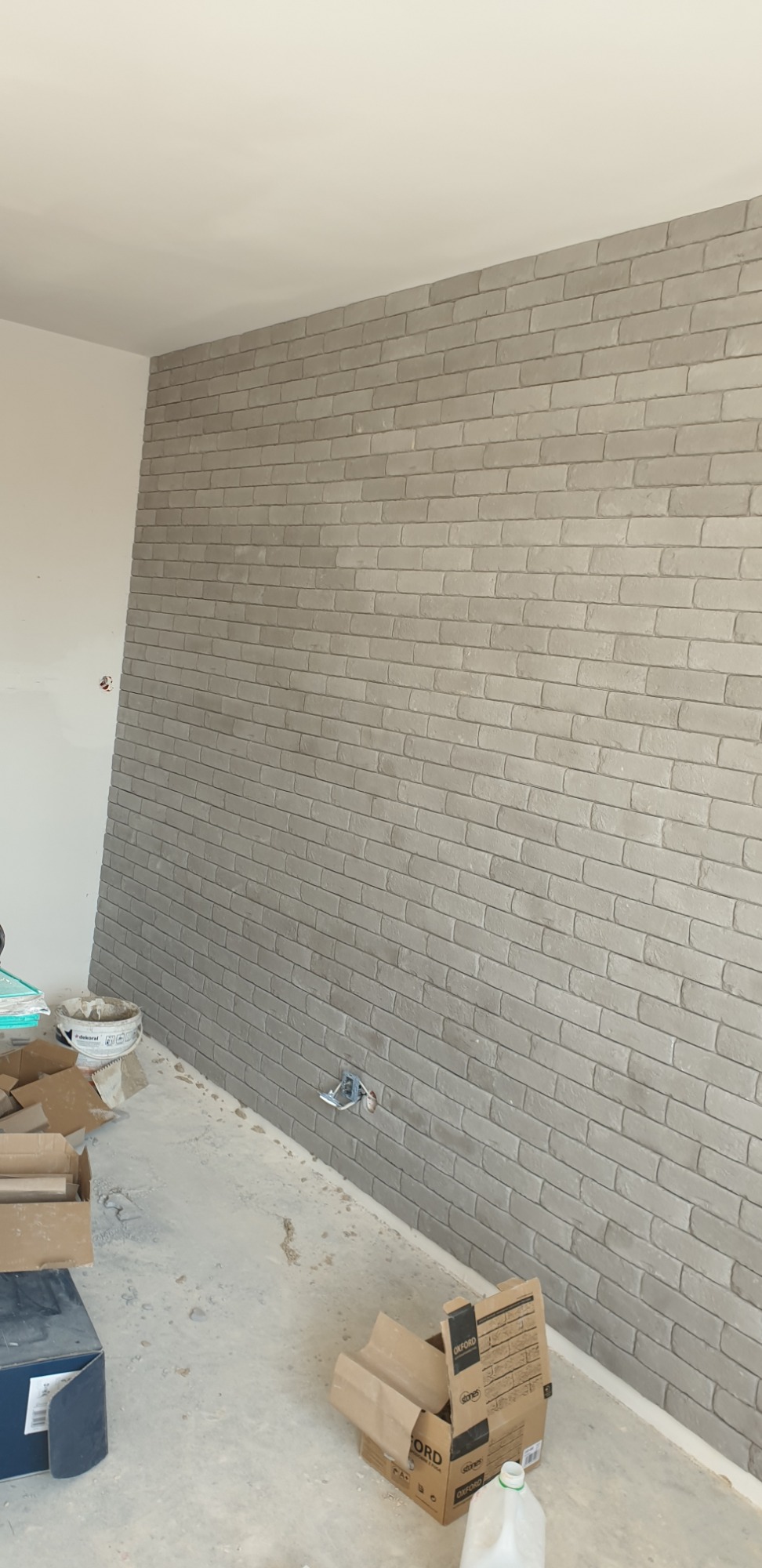 Ściana wykończona szarą cegłą dekoracyjną w trakcie remontu, widoczne puszki elektryczne, kartony i wiadro z zaprawą na podłodze.