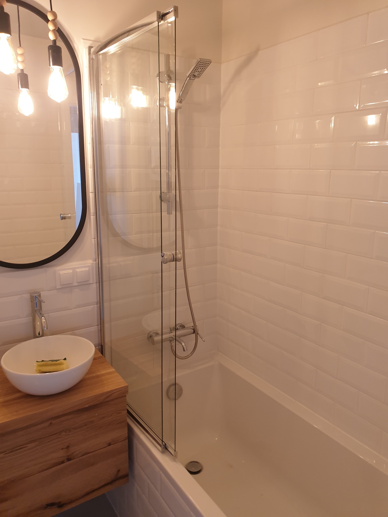 Nowoczesna łazienka z wanną za szklanym parawanem, białe kafelki typu metro, umywalka nablatowa na drewnianej szafce i okrągłe lustro z dekoracyjnym oświetleniem.