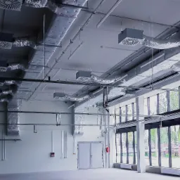 Instalacja wentylacyjna i rurociągi sprężonego powietrza pod sufitem hali produkcyjnej, widoczne kratki wentylacyjne, rury, kanały w izolacji, okna z widokiem na zewnątrz, drzwi i szafka...