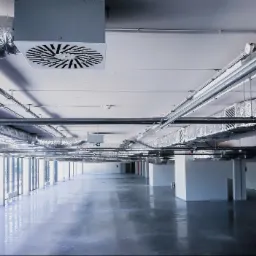 Hala produkcyjna z widoczną instalacją wentylacyjną pod sufitem, w tym kanały wentylacyjne owinięte folią aluminiową i okrągłe nawiewniki. Szara, polerowana posadzka odbija światło. Widoczne...