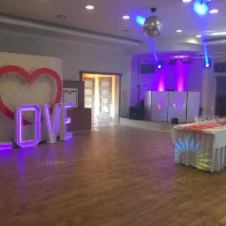 Sala bankietowa z dekoracjami: podświetlany napis LOVE, czerwone serce na tle tapety, parkiet, scena z nagłośnieniem podświetlona fioletowymi reflektorami, lustrzana kula i st&oacute;ł z kieliszkami.