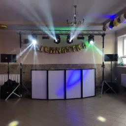 Sala przygotowana na imprezę: biały panel DJ-ski podświetlony na niebiesko, nagłośnienie po bokach, oświetlenie sceniczne z belką i reflektorami, złote balony foliowe z napisem 'WIEMNASTKA'...
