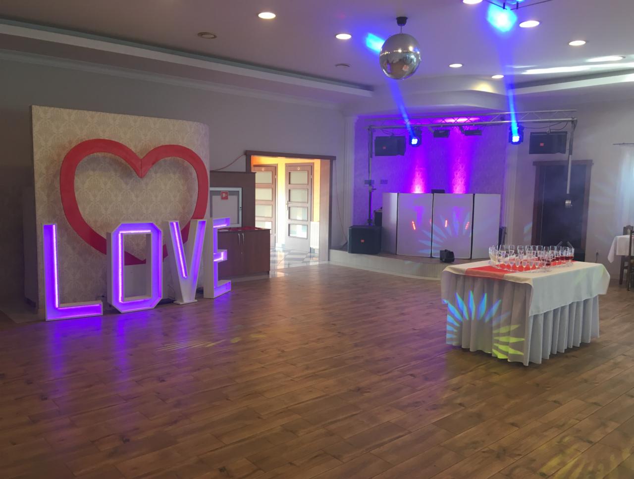 Sala bankietowa z dekoracjami: podświetlany napis LOVE, czerwone serce na tle tapety, parkiet, scena z nagłośnieniem podświetlona fioletowymi reflektorami, lustrzana kula i stół z kieliszkami.
