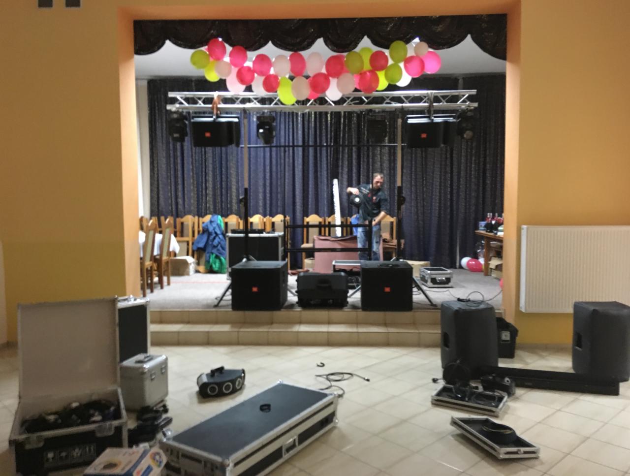 Sala bankietowa w trakcie przygotowań do imprezy: montaż nagłośnienia i oświetlenia scenicznego z kolorowymi balonami pod sufitem, widoczny mężczyzna regulujący reflektor.