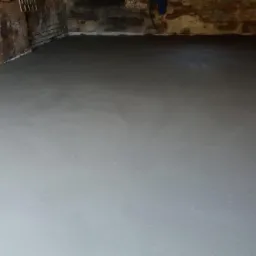 Świeżo wylana, gładka posadzka jastrychowa w pomieszczeniu z surowymi, ceglanymi ścianami, widoczne ślady wilgoci na murze.