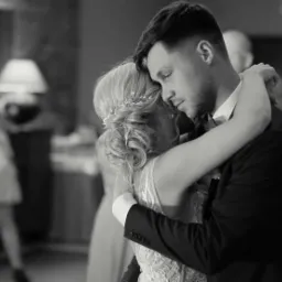 Czarno-biały kadr z filmu ślubnego: para młoda w objęciach podczas pierwszego tańca, widoczny fragment dekoracji sali i goście w tle.