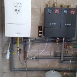 Biały piec gazowy Vaillant z podłączonymi rurami i dwoma szarymi modułami Bosch na ścianie z beżowych płytek.