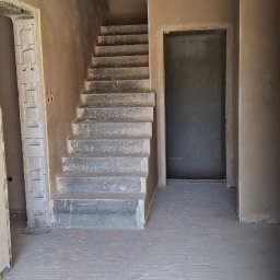 Piotr Ślubowski PIOTRBUD - Wnętrze w stanie surowym: surowe ściany, betonowe schody, żółte rury wentylacyjne pod sufitem. Widoczne ślady po tynkowaniu i szpachlowaniu. Wnętrze w trakcie prac wykończeniowych.