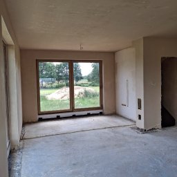 Piotr Ślubowski PIOTRBUD - Surowe tynki maszynowe w jasnym pokoju z dużym oknem na zewnątrz. Widoczne niedoskonałości i przygotowanie do dalszych prac wykończeniowych. W tle zielony krajobraz.