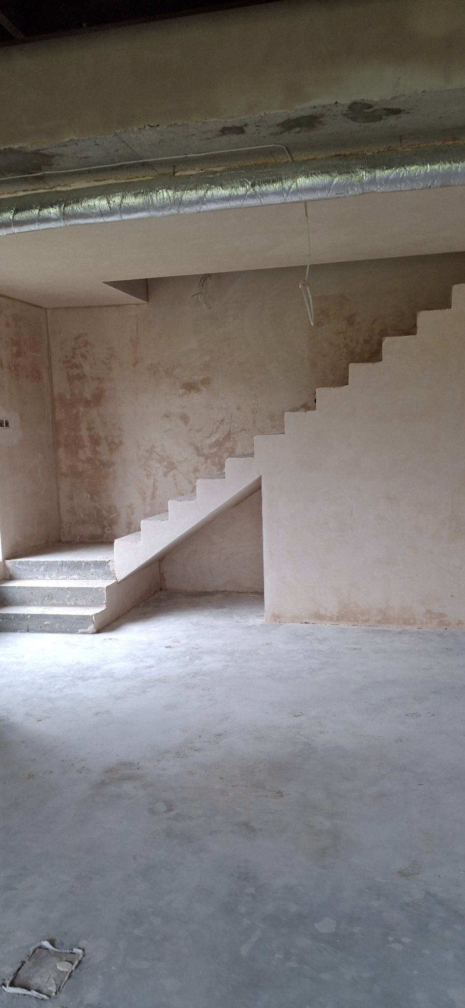 Surowe wnętrze po tynkowaniu: schody, ściany i sufit pokryte świeżym tynkiem. Widoczne niedoskonałości i faktura tynku. Metalowa rura wentylacyjna pod sufitem.