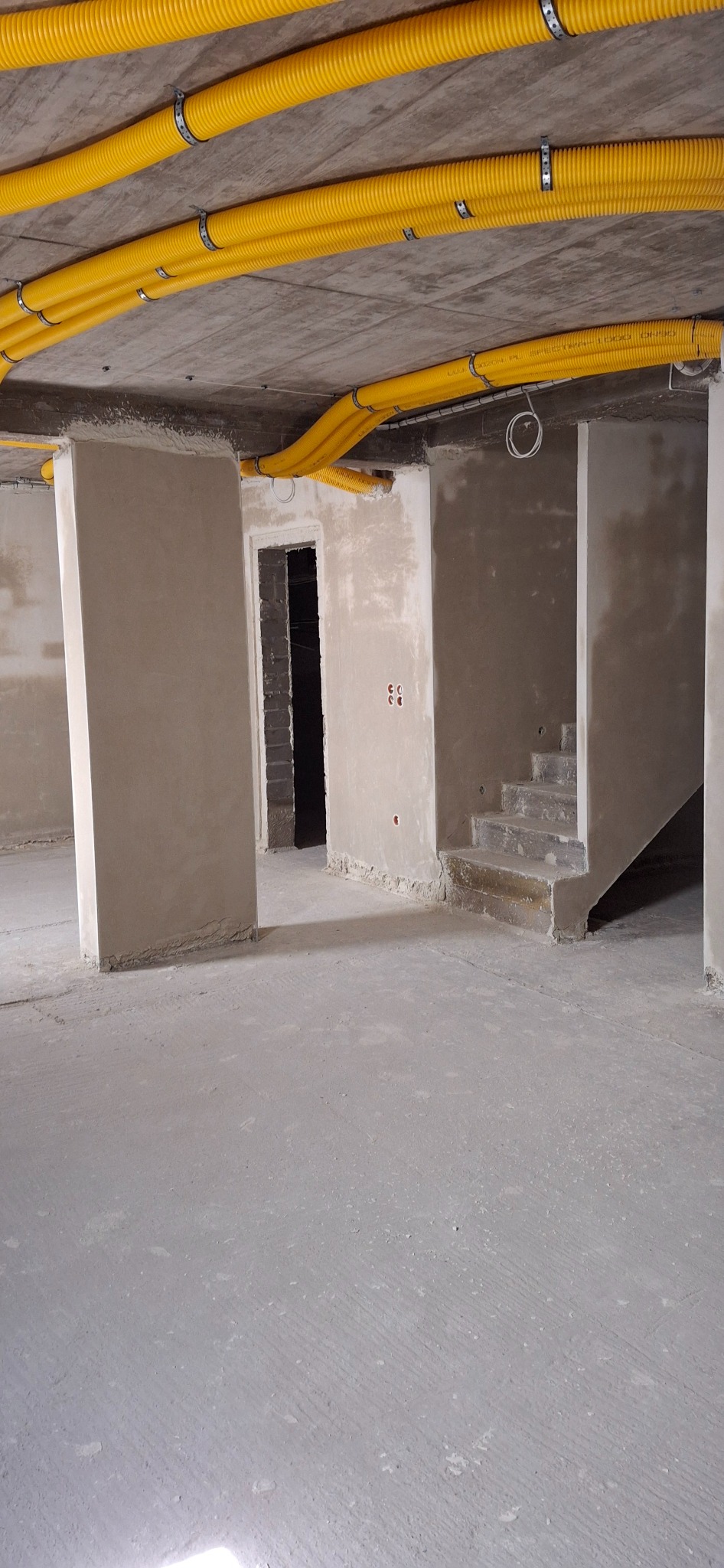 Surowy stan wnętrza: ściany po tynkowaniu, widoczne żółte rury wentylacyjne pod sufitem, betonowa podłoga i schody w trakcie wykończenia. Wnętrze w stanie deweloperskim.