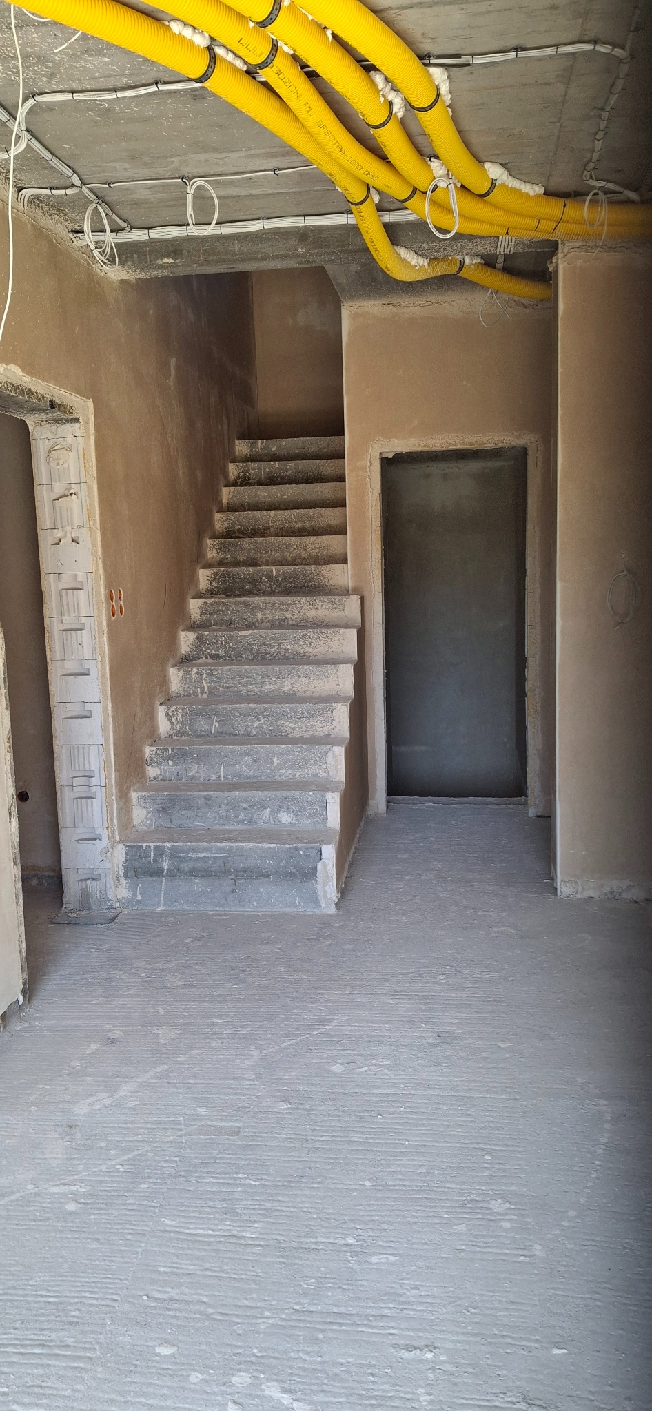 Wnętrze w stanie surowym: surowe ściany, betonowe schody, żółte rury wentylacyjne pod sufitem. Widoczne ślady po tynkowaniu i szpachlowaniu. Wnętrze w trakcie prac wykończeniowych.