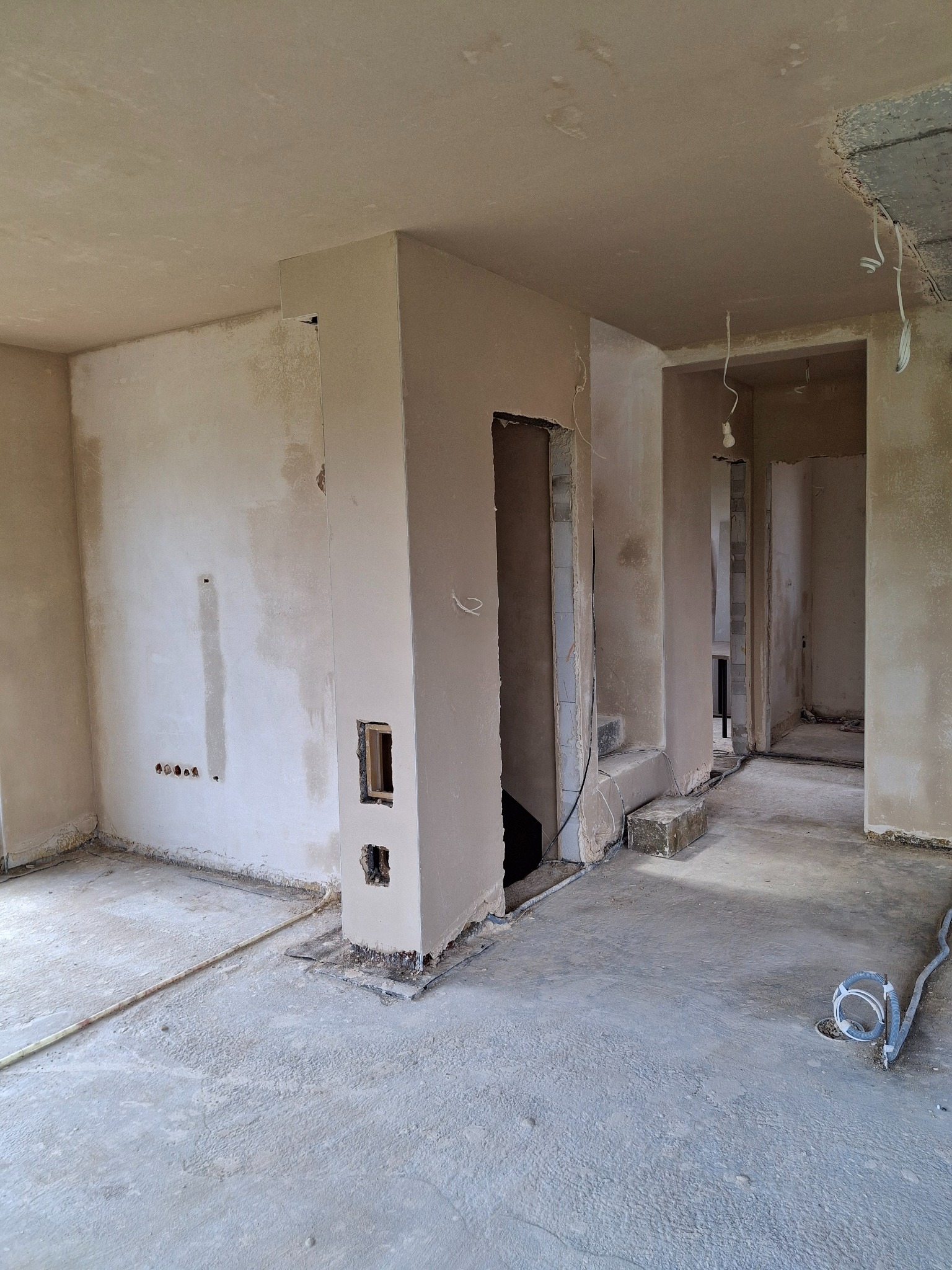 Surowe wnętrze w trakcie prac wykończeniowych: ściany pokryte tynkiem, widoczne instalacje elektryczne, surowy beton na podłodze, przygotowane otwory na drzwi.