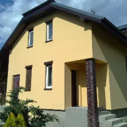 Żółta elewacja domu jednorodzinnego z brązowymi detalami: okna z roletami, kolumna wejściowa z cegły klinkierowej, widok z niskiej perspektywy na tle błękitnego nieba.