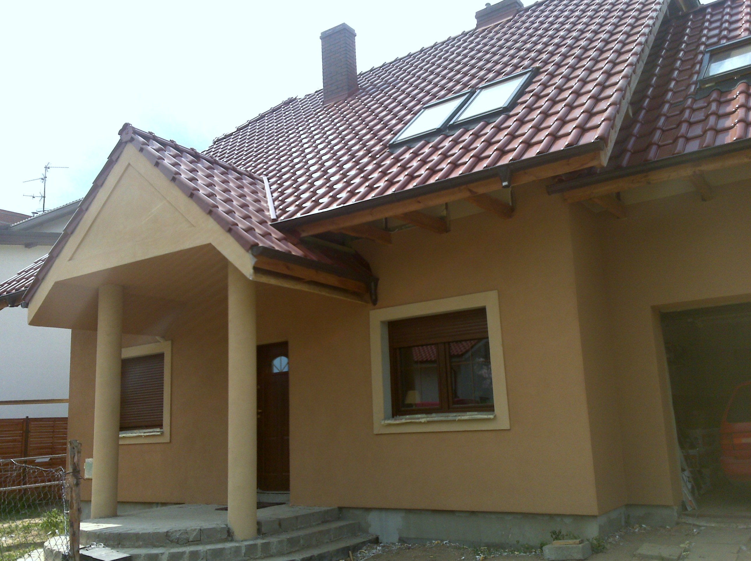 Dom jednorodzinny z nową elewacją w odcieniach beżu i brązu, widoczne okna połaciowe, okna z roletami zewnętrznymi i wejście z kolumnami.