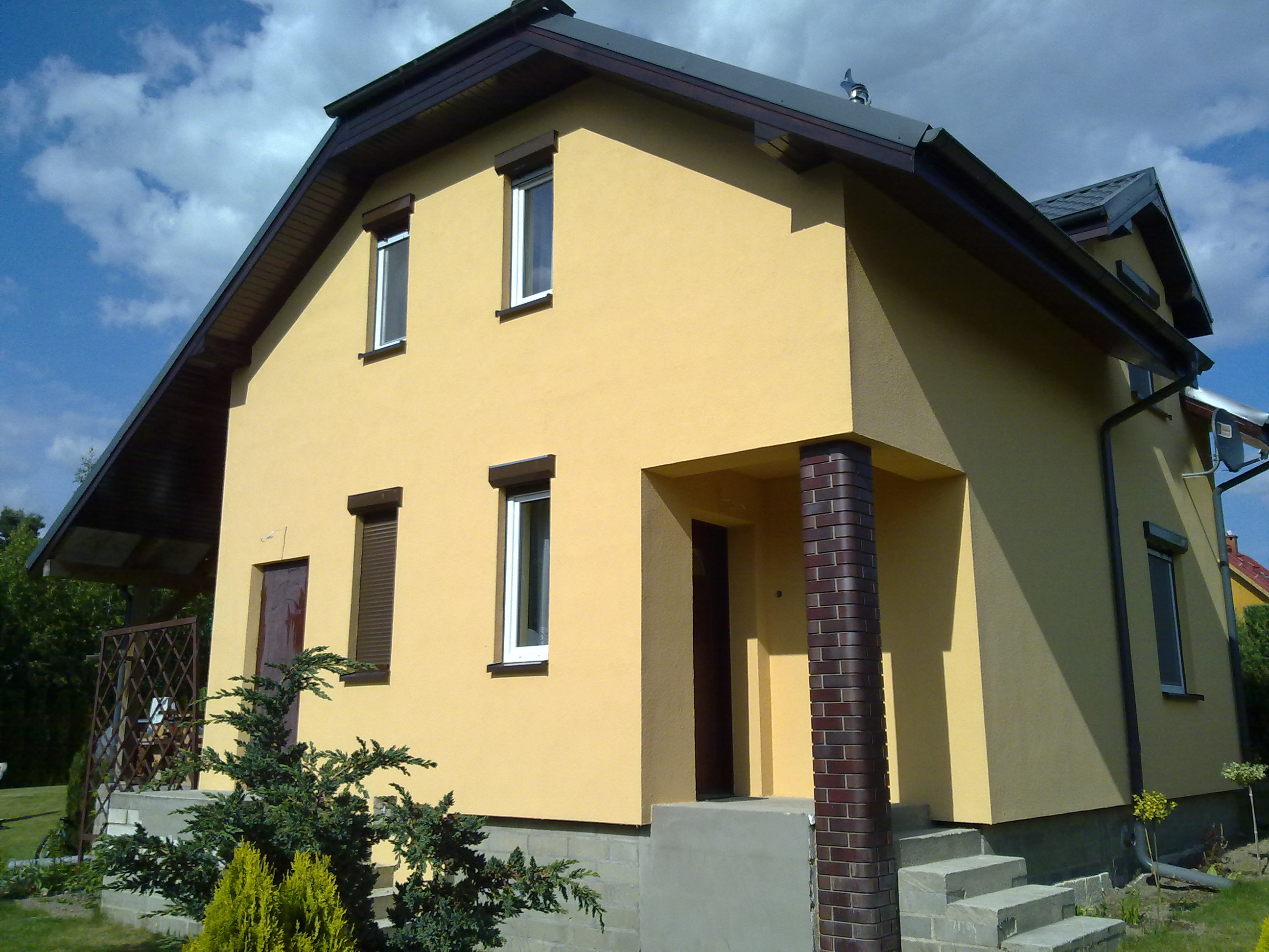 Żółta elewacja domu jednorodzinnego z brązowymi detalami: okna z roletami, kolumna wejściowa z cegły klinkierowej, widok z niskiej perspektywy na tle błękitnego nieba.