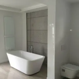 Nowoczesna łazienka z wolnostojącą białą wanną, grzejnikiem drabinkowym, płytkami imitującymi beton na ścianie i podwieszaną toaletą.