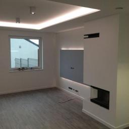 F.U.H. EArt - Nowoczesny salon z szarą podłogą imitującą drewno, białe ściany z podświetlanym panelem TV i minimalistycznym kominkiem, oświetlenie sufitowe LED.