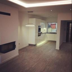 F.U.H. EArt - Jasne, przestronne wnętrze z drewnopodobną podłogą, białymi ścianami i nowoczesną kuchnią z czarnym blatem, oświetleniem LED podszafkowym i wiszącą lampą, widok na kominek i okno z zielenią...