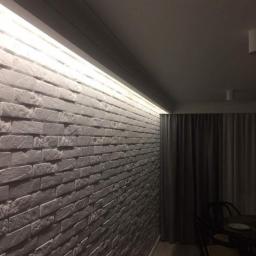 F.U.H. EArt - Ściana z szarej cegły dekoracyjnej z podświetleniem LED wzdłuż sufitu, widok z dołu pod kątem, zasłonięte okno w tle.