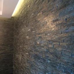 F.U.H. EArt - Ściana wyłożona ciemnym, błyszczącym kamieniem łupkowym ułożonym w poziomych pasach, oświetlona od góry listwą LED, tworząca surowy, ale elegancki efekt.