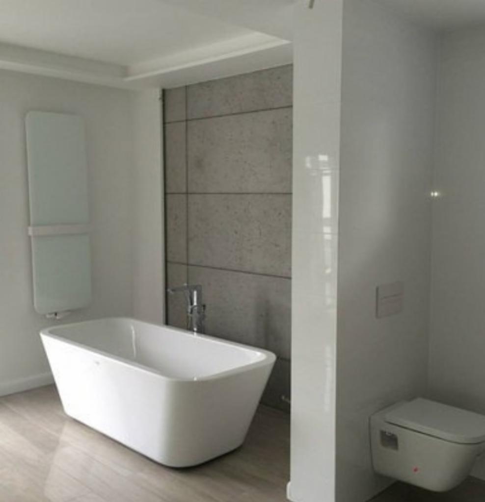 Nowoczesna łazienka z wolnostojącą białą wanną, grzejnikiem drabinkowym, płytkami imitującymi beton na ścianie i podwieszaną toaletą.