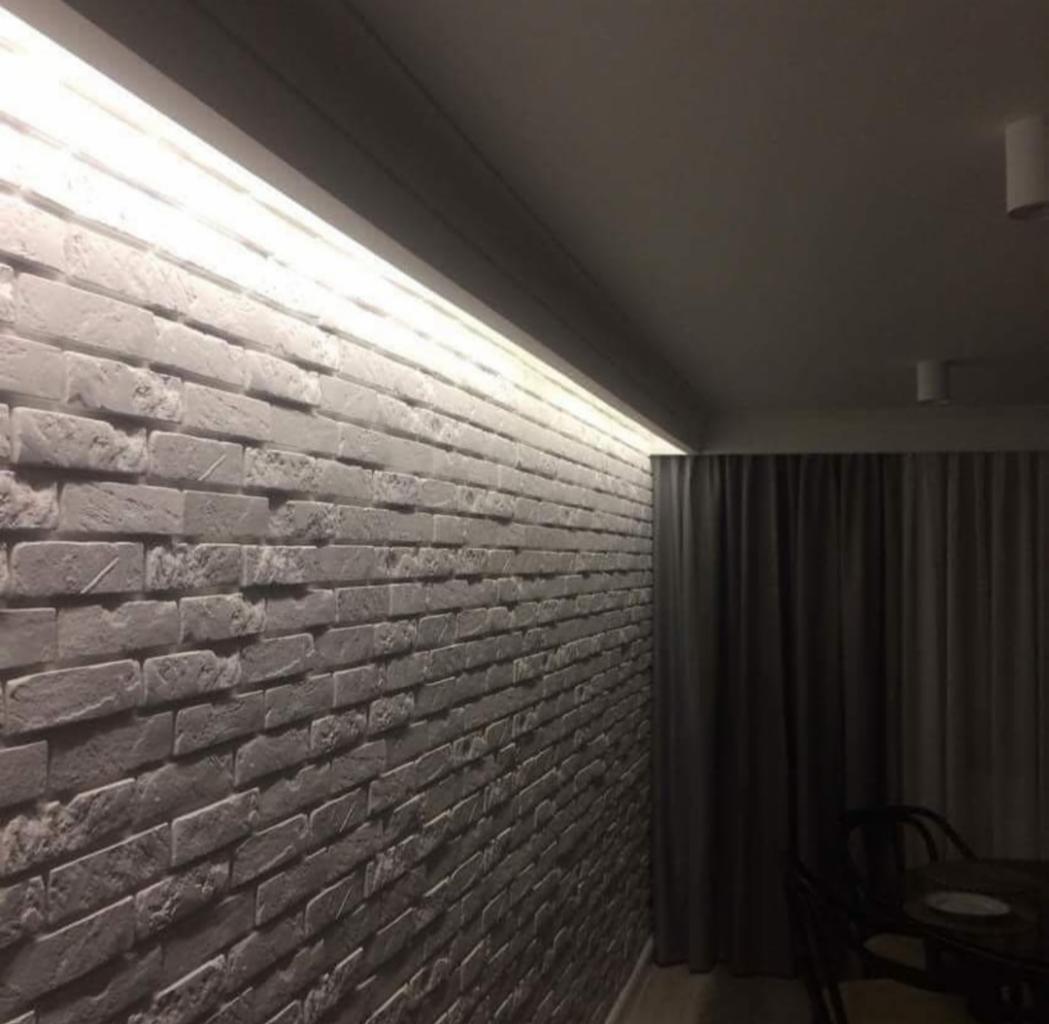 Ściana z szarej cegły dekoracyjnej z podświetleniem LED wzdłuż sufitu, widok z dołu pod kątem, zasłonięte okno w tle.
