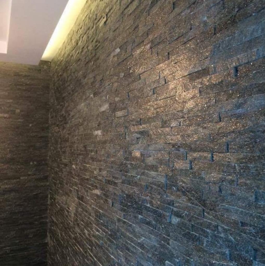 Ściana wyłożona ciemnym, błyszczącym kamieniem łupkowym ułożonym w poziomych pasach, oświetlona od góry listwą LED, tworząca surowy, ale elegancki efekt.