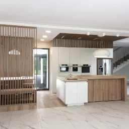 Nowoczesna kuchnia z wyspą o drewnianym wykończeniu, białe szafki, drewniana ażurowa ścianka działowa i marmurowe płytki podłogowe w przestronnym wnętrzu.