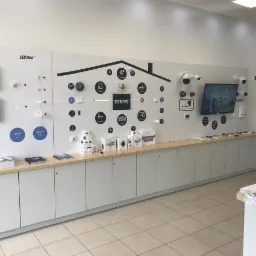 Ekspozycja systemów inteligentnego domu i monitoringu w salonie, prezentująca panele sterowania, kamery, czujniki i oświetlenie LED, z elementami graficznymi symbolizującymi funkcje, oraz logo...