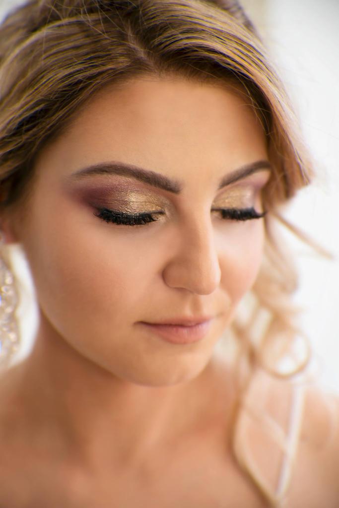 Portret kobiety z zamkniętymi oczami, prezentujący makijaż oczu z użyciem złotych i bordowych cieni oraz sztucznych rzęs, z delikatnym makijażem ust i blond włosami upiętymi w loki.