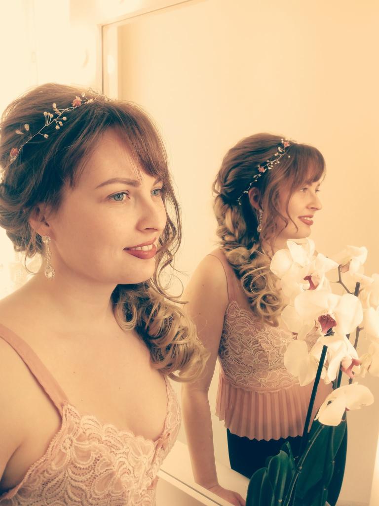 Kobieta z upiętymi włosami, ozdobną opaską i kolczykami, odbijająca się w lustrze, trzymająca białe orchidee, w różowej koronkowej bluzce.