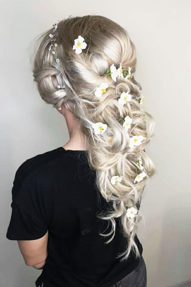 Upięcie ślubne z jasnych włosów z wplecionymi białymi kwiatami i ozdobną spinką, widok z tyłu na tle jasnej ściany.