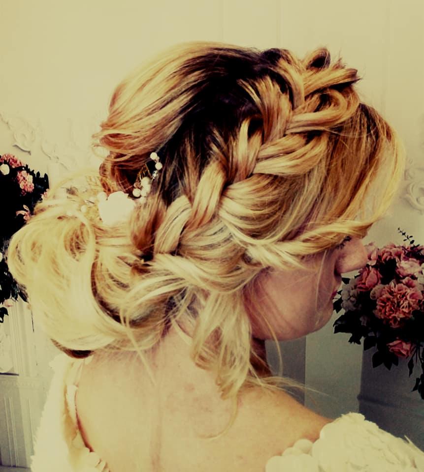 Upięcie ślubne z warkoczem, blond włosy z ciemnymi odrostami, ozdobione perłowymi spinkami, widok z tyłu, bukiety kwiatów w tle.