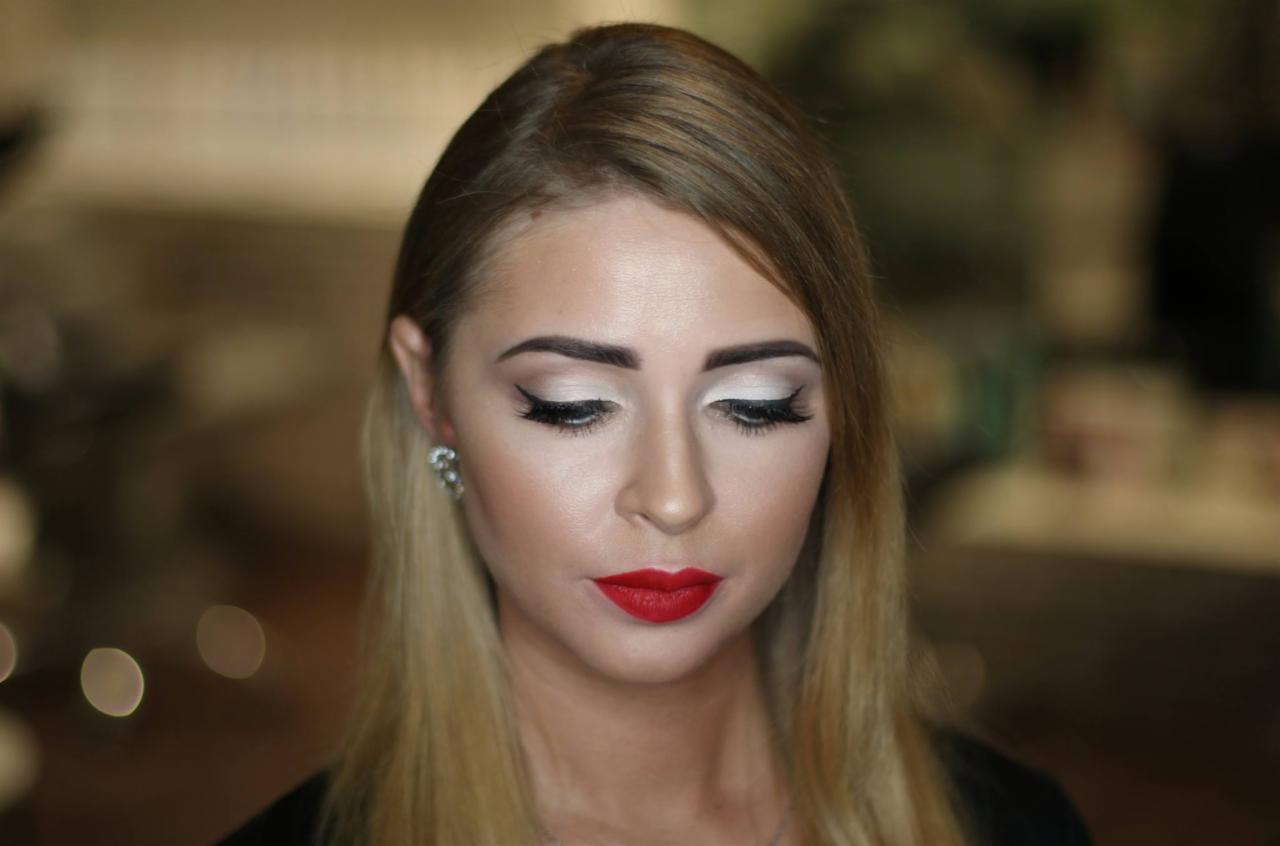 Portret kobiety z blond włosami, w pełnym makijażu: białe cienie, czarny eyeliner i intensywnie czerwone usta, z delikatnymi kolczykami w uszach.