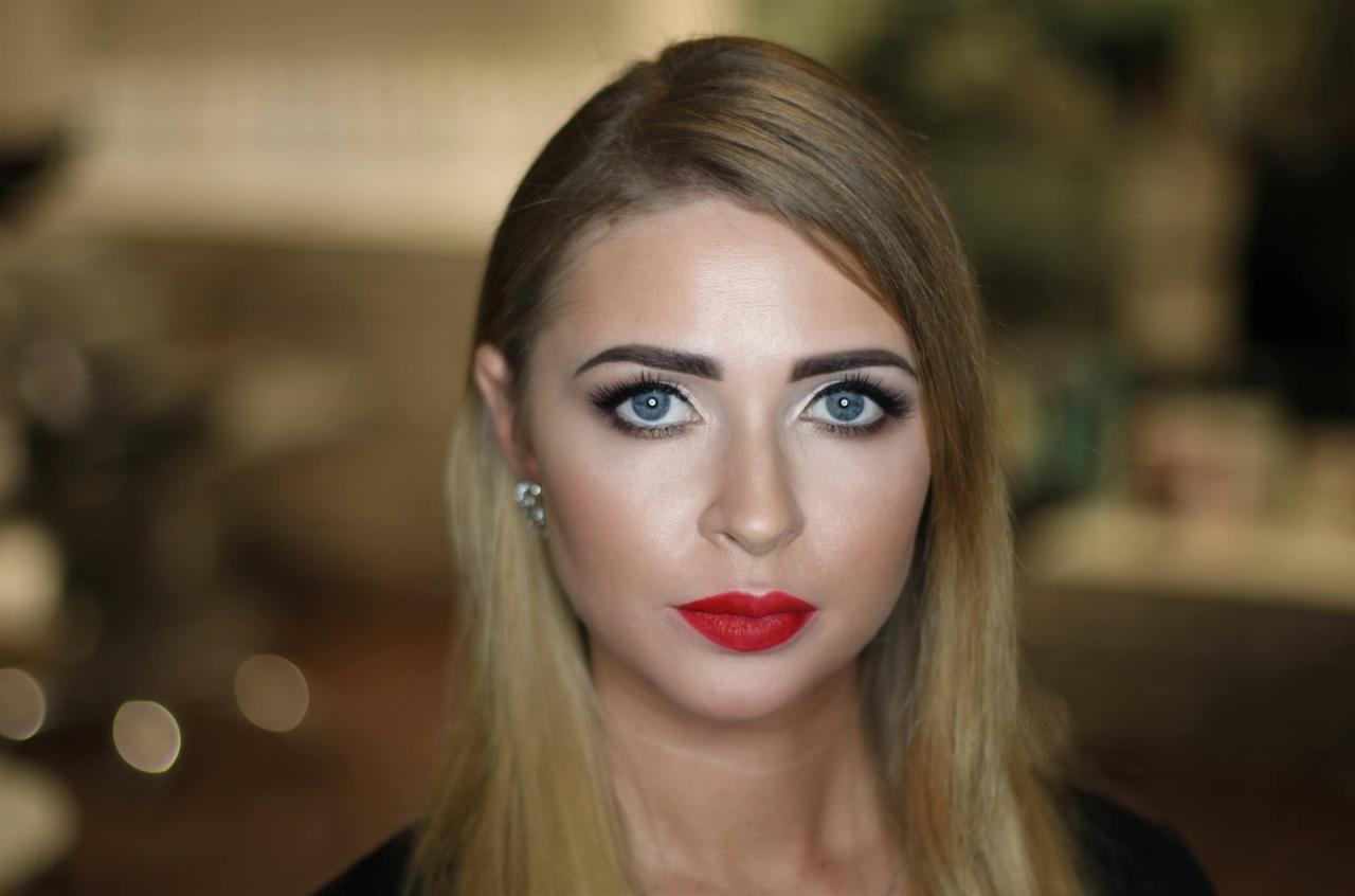 Portret kobiety z profesjonalnym makijażem, mocno podkreślone oczy i czerwone usta, widoczne kolczyki z kryształkami, rozmyte tło.