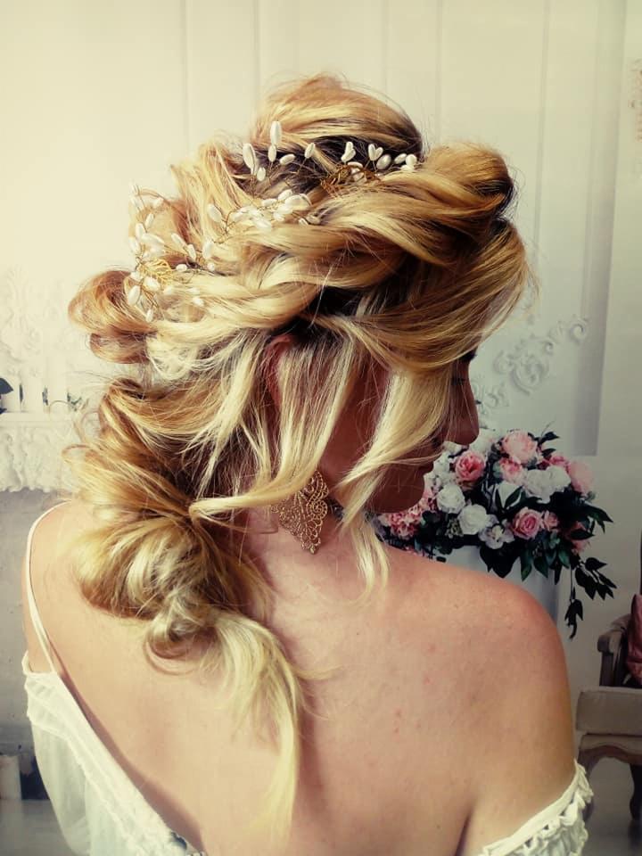 Upięcie ślubne z blond pasemkami, ozdobione delikatną biżuterią z perełek, widoczne złote kolczyki i kwiaty w tle.